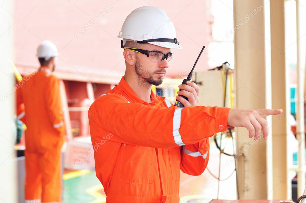 An experienced sailor in orange overalls and a white helmet speaks on a walkie-talkie and gives instructions to other workers.