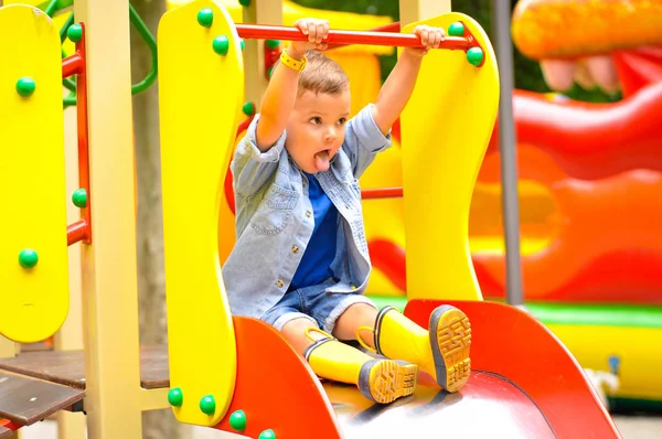 A little cute three-year-old boy plays on the playground and slides down the slides in yellow rubber boots.