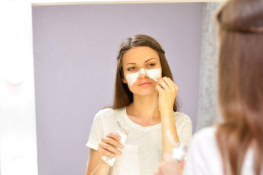 Genç bir güzel kadın beyaz bir t shirt soyunma odası aynaönünde evde dururken onu temizlenmiş yüzüne beyaz bir maske koyar.