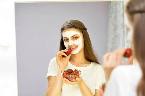 Young beautiful woman with a cosmetology mask on her face eats strawberries and looks in the dressing mirror