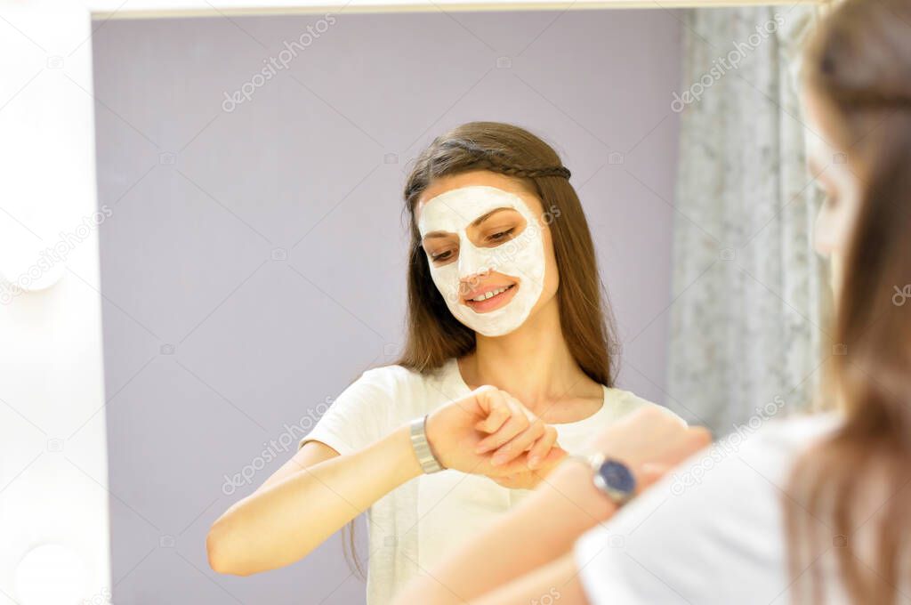 A young beautiful woman puts on a white mask on her cleansed face while standing at home in front of the dressing room mirror in a white T shirt.