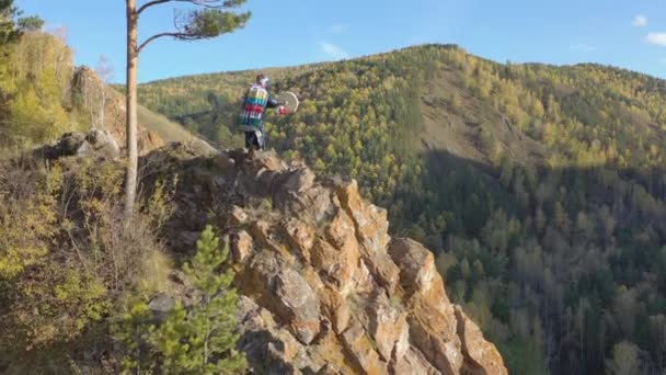 Luchtfoto van een oude tovenaar in een helder sjamaans kostuum dansend met een tamboerijn op een bergtop. — Stockvideo