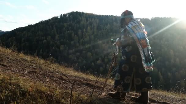 Egy öreg sámán fényes ruhában felmászik a hegyre egy erdei ösvényen.. — Stock videók