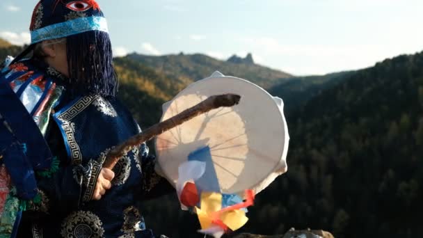 Un viejo chamán con un brillante traje nacional entra en trance golpeando una pandereta en la cima de una montaña . — Vídeo de stock