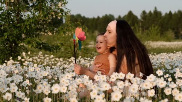 Медленное движение молодой привлекательной матери с сыном, играющим на ветряной мельнице, сидящей на цветочном поле на закате . — стоковое видео