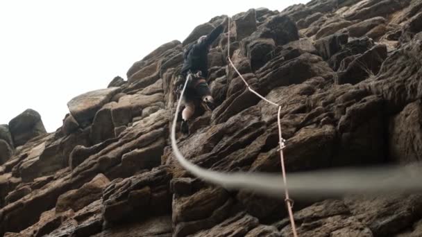 En manlig klättrare försäkrar ett rep klättra en stenig rutt i regnet. — Stockvideo