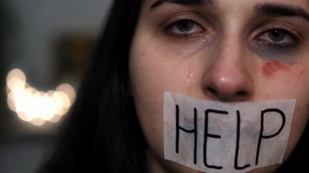 Slow motion portrait of a young woman victim of domestic violence, the mouth is sealed with a white plaster with the inscriptionon HELP, tears running from her eyes. — Stock Video