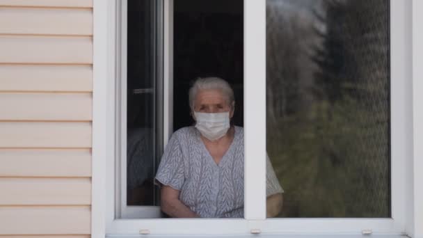 Sorgsen äldre kvinna i en medicinsk mask står hemma vid fönstret och titta på gatan. — Stockvideo