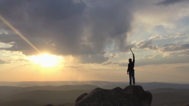 Drone shot van een silhouet van een bergbeklimmer die op de top van een berg staat en overwinnend zijn hand optilt met een ijsbijl bij zonsondergang. — Stockvideo