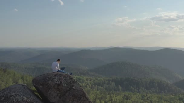 森の中の岩の山の上に座っているラップトップで働いている若い男の空中ビュー. — ストック動画