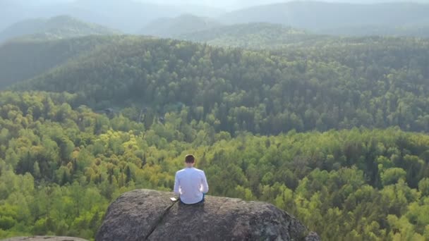 絶壁の上の岩の山の端に座っているオープンエアのラップトップで働いているフリーランスのドローン映像. — ストック動画