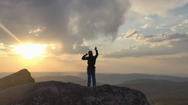 Drone schot van een man die op een rots berg staat en vrolijk zwaait met zijn handen met een ijsbijl bij zonsondergang. — Stockvideo