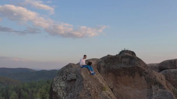 森の中の山の頂上にある岩棚の端に座っているラップトップで働いている若い男のドローンショット. — ストック動画