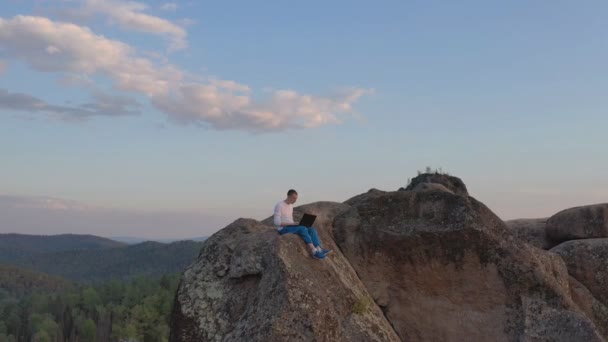 Drone disparo de un joven trabajando en un portátil sentado en el borde de una cornisa de roca en la cima de una montaña en el bosque . — Vídeo de stock