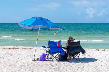 Yaşlı bir kadın, deniz ve gökyüzünün arka planında, bir plaj şemsiyesinin gölgesinde oturur, bulutlar, plaj, güzel bir yaz sabahı, okyanusta kocasına bakar. Bell Air Plajı, Florida, Usa
