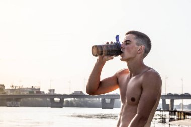 Mavi havluyla genç erkek sporcu şişeden su içiyor.