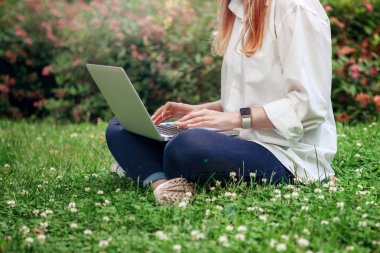 Parktaki çimenlikteki kız dizüstü bilgisayarla çalışıyor. Yeşil çimlerin üzerinde oturan dizüstü bilgisayarı olan mutlu kadın. Uzaktan öğrenme ve uzaktan çalışma kavramı