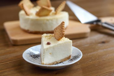 Klasik bir New York peynirli keki tabakta kare kurabiyelerle süslenmiş, kahve dükkanındaki ahşap masanın üzerinde, arka planda bütün pastayla birlikte. Tatil için tatlı pasta, doğum günü partisi