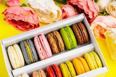 Sarı renkte romantik bir doğum günü kutlaması için hediye kutundaki renkli badem kurabiyeleri, pion çiçekleri, saten kurdele üst görünümü, birçok farklı türde makaron çeşitleri elle süslenmiş altın gümüşler.