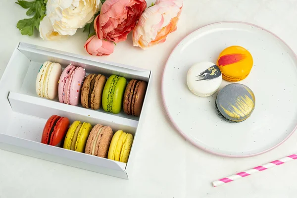 colorful modern macaroons in gift box and on plate for celebration romantic party birthday on white, pion flowers, satin ribbon top view, many different types flavors of macarons hand drawn decorated golden silver