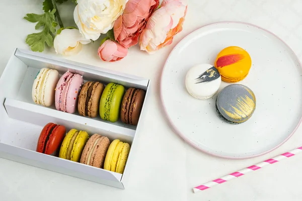 colorful modern macaroons in gift box and on plate for celebration romantic party birthday on white, pion flowers, satin ribbon top view, many different types flavors of macarons hand drawn decorated golden silver