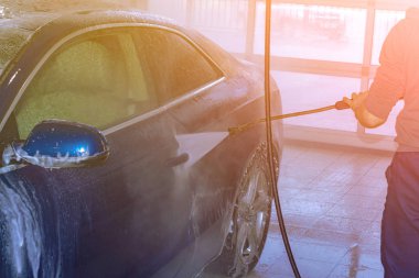 Bir oto yıkama işçisi yüksek basınçlı bir aparatla mavi bir arabayı yıkar. Köpük. Arka kenar görünümü.