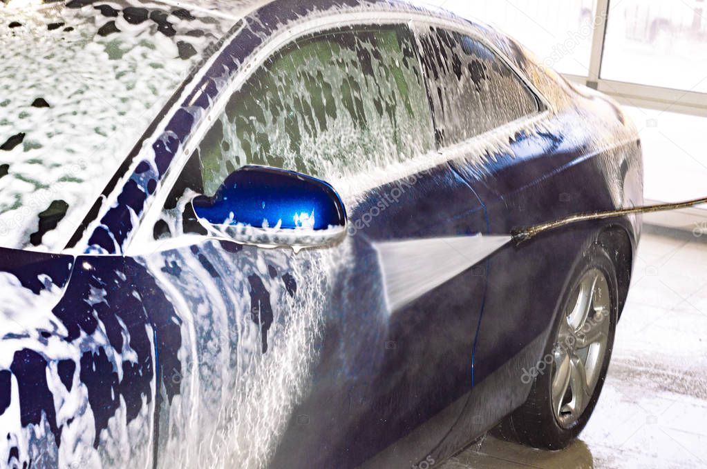 A car wash worker washes blue car with high-pressure apparatus. Foam. Rear side view.