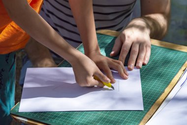 Çocuk ve yetişkin parkta. Hild bir objeyi beyaz kağıt üzerinde bıçakla keserken bir yetişkin ona yardım ediyor. Yan görünüm.