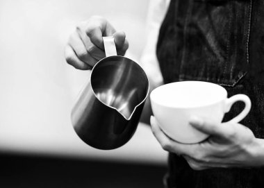 Barista bir kafede çalışıyor, Barista kahveye süt koyuyor, Barista da latte sanatıyla kahve yapıyor.