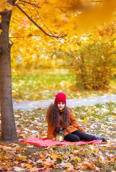 Beautiful sad teenage girl in a red hat and an orange jacket sits under a tree with a lantern. The yellow maple leaves on the trees in the background are blurred. Autumn vertical photo of a child