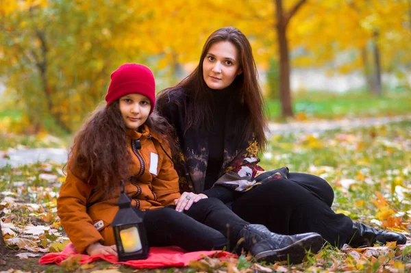 Beautiful mom and daughter teenager on a picnic in autumn. A child in a red hat and an orange jacket, a woman in black clothes. Sitting under a tree on a plaid. In the background are yellow maple leav