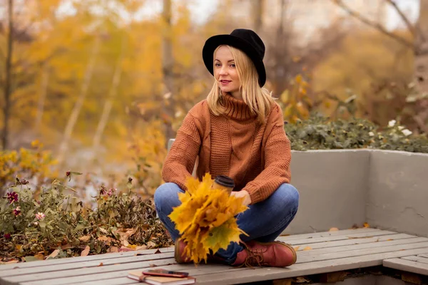 relaxed young blonde in a brown warm sweater, black felt hat, blue jeans and boots sits on a bench in the autumn in the park, holds a bouquet of leaves, looks forward