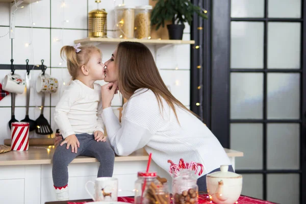 Mom kisses her little daughter in white sweaters in a festive br