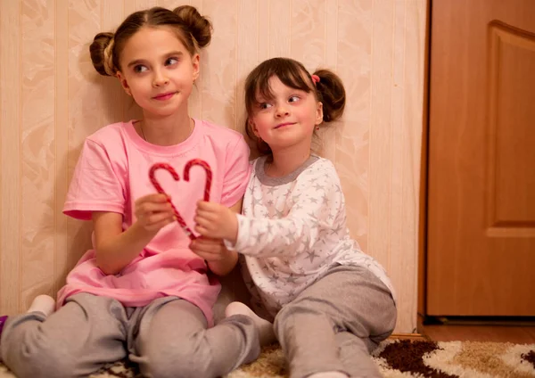 kids pajama party. funny girls in pink, white and gray pajamas with bob hairstyles play with red christmas candy canes, make a heart.