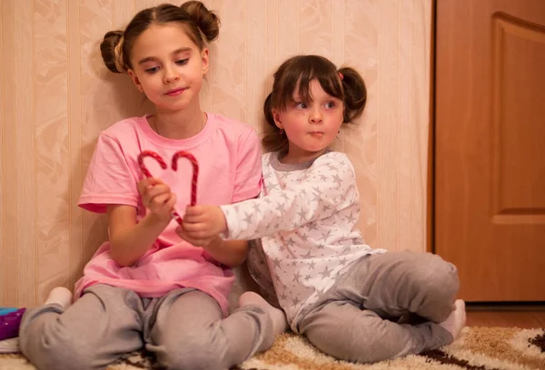 kids pajama party. girls in pink, white and gray pajamas with hairstyles in bunches sit on the carpet and play with red Christmas candy canes.