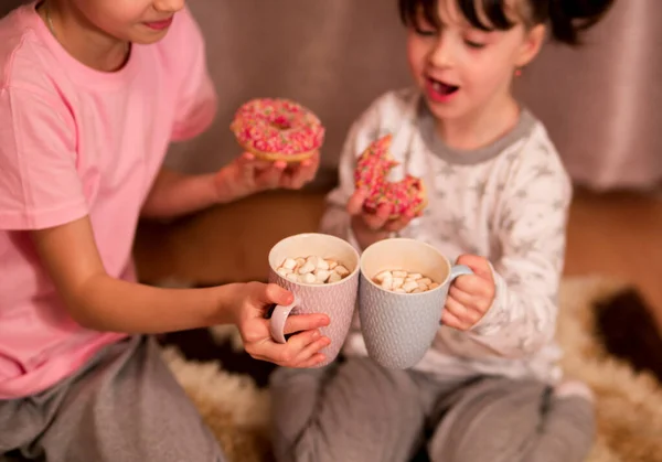 children\'s pajama party. funny girls in pink, white and gray pajamas with bob hairstyles eat donuts and drink from a cup of cocoa with marshmallows