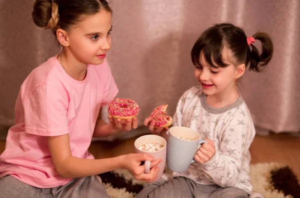 children\'s pajama party. funny girls in pink, white and gray pajamas with bob hairstyles eat donuts and drink from a cup of cocoa with marshmallows.
