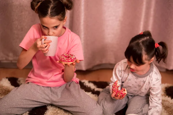 children\'s pajama party. sisters in pink, white and gray pajamas with bob hairstyles eat donuts and drink from a cup of cocoa with marshmallows