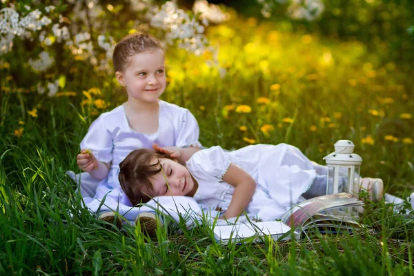 two girl friends in white dresses are sitting on the grass in a blooming cherry garden. one of them is sleeping on the lap of the other.