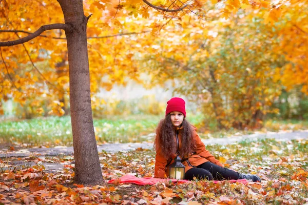 Beautiful teenager girl in a red hat and an orange jacket sits on a plaid with a lantern. Autumn landscape. The yellow maple leaves on the trees in the background are blurred