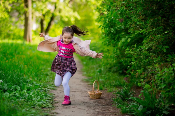 cheerful child girl with a tail in a pink coat, plaid dress and boots runs forward along the path. around beautiful bushes, grass.