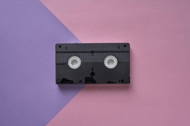 Pembe ve mor arkaplan üzerine siyah VHS kasetin üst görüntüsü. Minimalist retro konsept.