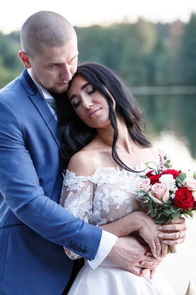 Pareja en traje de boda en el fondo del lago al atardecer, la novia y el novio. Sonrían el uno al otro. — Foto de Stock