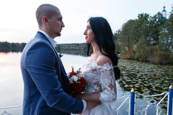 La novia y el novio sonríen y se miran. La novia sostiene un ramo. Paseo de bodas en el pueblo . — Foto de Stock