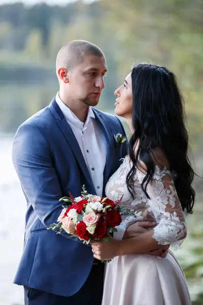 La novia y el novio sonríen y se miran. La novia sostiene un ramo. Paseo de bodas en el pueblo . — Foto de Stock