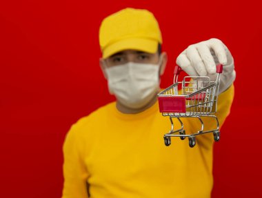 Tıbbi kauçuk eldiven ve maskeli bir sepet taşıyan teslimatçı. Uzayı kopyala Hızlı ve ücretsiz nakliye. Çevrimiçi alışveriş ve hızlı teslimat. Karantinaya alındı. Koronavirüs. Objenin kendisi odak dışı. kırmızı arkaplan