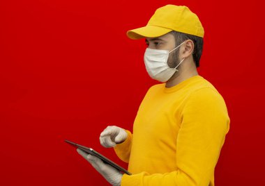 Sarı şapkalı teslimatçı, steril yüz maskesi eldivenli boş tişörtlü kırmızı arka planda izole edilmiş bir tablet tutuyor. Çalışan kurye servisi karantina virüsü koronavirüs konsepti. Dümdüz, ayakta duruyor le