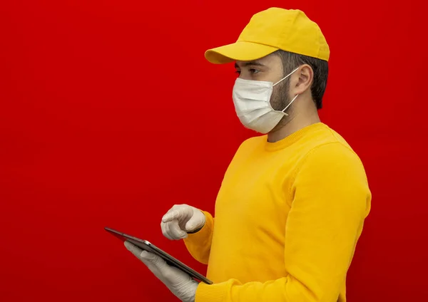 黄色の帽子の配達人 滅菌顔のマスク手袋の形をした空白のTシャツ 赤い背景に隔離されたタブレットを持っています 従業員の作業宅配便サービス隔離パンデミックウイルスコロナウイルスの概念 真っ直ぐに見ると — ストック写真