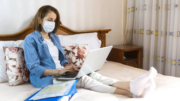 A woman in a medical mask works remotely from home on the bed. Using a computer. Distance learning online education and work. using a medical mask