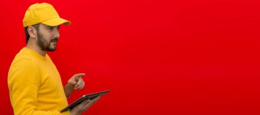 Sarı şapkalı, boş tişörtlü, elinde tablet olan, kırmızı arka planda izole edilmiş bir teslimatçı. Kurye servisi çalışanı. Logo ve mesaj için yer. Yerleşim planı. Model